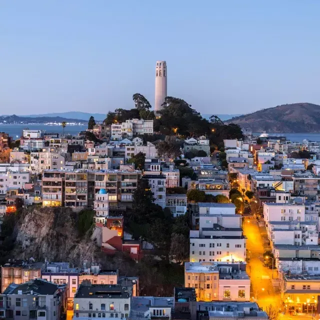 La Coit Tower de 贝博体彩app au crépuscule, avec des rues éclairées devant elle et la baie de 贝博体彩app derrière elle.